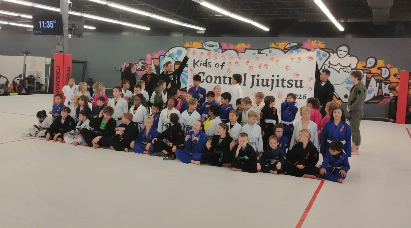 The whole kids program sitting together on the mat in front of the Control Jiu-Jitsu mural